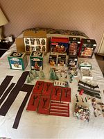 Wide view on bed showing multiple boxed and unboxed Holiday Village collectible porcelain houses, miniature trees, figurines, track pieces, and accessories laid out