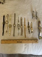 Overview of all 13 ladies wristwatches laid out on white fabric with ruler for scale showing varying band types and face shapes/colors.