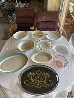 Top-down photo of all serving dishes and trays on a white tablecloth with two brown chairs in the background. Shows a variety of dishes including white ceramic, glass, and a dark decorative tray.