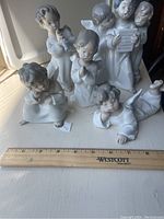 Seven porcelain angel figurines displayed with a ruler for scale showing approximately 4 to 6 inches tall. Figurines include an angel playing flute, praying angel, a group reading sheet music, and a reclining angel.