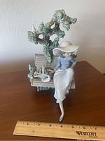 Wide view showing woman seated on bench, tree background, basket of flowers, and a white bird.