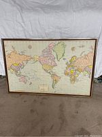 Framed world map resting on carpet, showing front side with detailed country borders and pastel color coding.