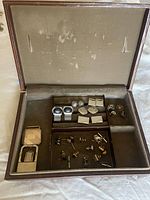Open men's jewelry case showing five pairs of cufflinks, three tie tacks, three pins, and two belt buckles arranged inside compartments.