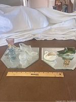 Overview of two mirror trays each holding glass and ceramic miniature figurines including shoes, swan, bottles, and goblet shape.