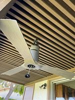 Photo showing the modern ceiling fan mounted on an outdoor patio ceiling, three wide light wood finish blades attached to a cylindrical matte grey motor housing with wiring exposed.