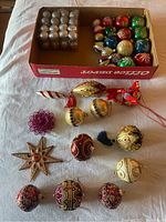 Box of assorted mini silver and colorful glass Christmas ball ornaments along with loose assorted shaped and colored ornaments spread on white background.