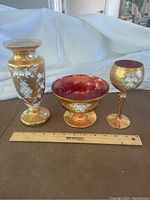 Set of three Murano glass items on brown surface with ruler for scale, showing vase, footed bowl, and goblet decorated with gold and flowers.