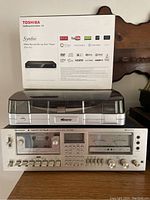 Photo showing Toshiba Symbio Media Box sealed box placed behind Memorex USB turntable and Sharp RT-3388 Stereo Cassette Deck, arranged stacked on a wooden surface.