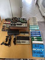 Overview of all items on table: Sony reel-to-reel recorder with two reels engaged, assorted reel-to-reel tapes in sleeves arranged to the side, three microphones including boxed Sony F-96 and two others, manual booklet.