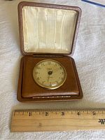 Vintage Gilbert travel alarm clock face and case open, showing dial and leatherette with satin interior lining, ruler for scale.