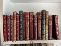 Photo showing 15 miscellaneous pocket size books mostly in red leather bindings with gold embossed titles and designs, arranged upright on a shelf.