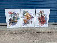 Three stretched canvas art panels leaning against blue metal door outdoors on concrete. Each canvas 24x18 inches featuring illustrations of a trombone, French horn, and violin on sheet music backgrounds.