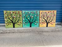Three square canvas paintings side by side showing stylized trees with black branches and multicolored circular leaf shapes on colored backgrounds blurry dust visible