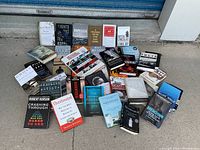 Photo of 27 used books arranged on ground showing front covers with some titles legible and visible signs of wear like tears and folds.