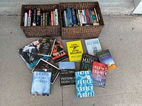 Two woven baskets filled with paperback books, with additional books laid out on the ground in front showing titles and covers.
