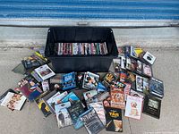 Photo showing a large black storage bin filled with DVDs and a wide spread of DVD cases around it on a concrete floor.