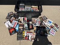 Wide shot showing a large black container filled with DVDs and many DVD cases arranged around it on the ground.