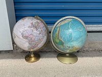 Two world globes side by side showing the full lot with metal bases on concrete ground in front of a blue door.