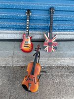 Full view of two miniature electric guitars and one miniature violin against a concrete and blue metal background.