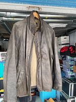 Brown leather jacket hanging on wooden hanger. Front view showing zipper, snap button closure, two front pockets and beige lining.