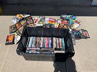 A large black bin filled with DVDs with more DVDs laid out behind it on the ground showing variety of titles.
