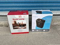 Photo of the desk lamp and Omnitech shredder boxes placed on concrete in front of a blue metal door, showing box front designs and item names.