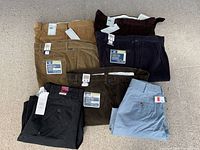 Seven assorted men's pants and shorts folded on the floor, showing tags and size labels 38/30, in various colors including brown, dark brown, navy blue, black, light blue, and khaki.