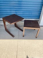 Two patio tables shown side by side on concrete ground with blue metal wall behind. One table features a curved bronzed metal base and wood slat top, the other has a beige metal base and wood slat top. Used condition with visible scratches and dust.