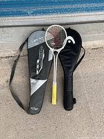 Photo showing a pink badminton racket with yellow grip between two black carrying cases, one Dunlop-branded, on concrete floor under blue metal door.