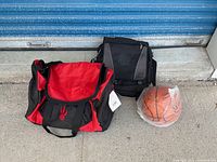 Full lot view showing two Raptors-branded bags and a basketball on concrete surface against a blue metal backdrop