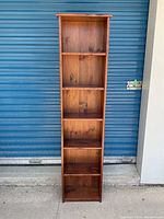 Front view of a tall, narrow wooden shelving unit with five open shelves, placed outdoors against a blue metal door.
