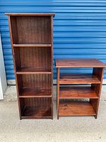Photo showing the front view of the two wooden shelving units side by side outdoor on pavement in front of a blue roll-up door. The taller unit has four shelves, the shorter has three shelves.