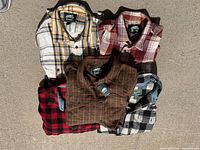 Top-down view of five folded Roots shirt jackets in various plaid patterns and colors laid out on a concrete floor.