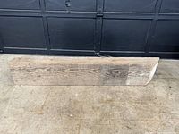Photo showing the full antique barn beam on a concrete surface in front of a dark garage door, highlighting its weathered texture and rectangular shape.