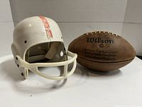 Photo showing vintage white football helmet with red stripe and attached facemask next to a Wilson branded NFL leather football, both showing wear.