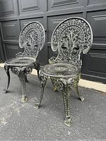 Front angled view of two ornate metal cafe chairs showing detailed backs and legs with worn paint.
