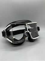 Side view of goggles showing chrome frame, clear glass lenses, and black leather padding with elastic strap attached.