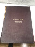 Closed dark brown book with gold embossed text 'CORONATION CEREMONY'. Visible wear and some damage to spine.