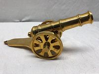 Side view of the small brass decorative cannon figurine showing polished cylindrical barrel and spoked wheels with attached carriage base.