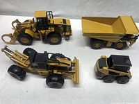 Four yellow CAT heavy machinery toy trucks including a loader, wheeled scraper, dump truck, and skid steer displayed on white surface.