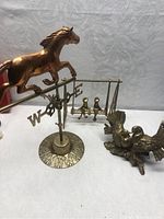 Side view of the brass horse weather vane showing directional letters and decorative base alongside brass swing set and eagle figurine.