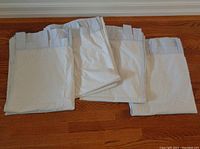 Four folded fabric curtains laid flat showing loops for hanging on a rod; light blue striped pattern visible under folds.