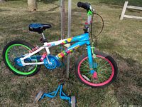 Full side view of the child's bicycle showing multicolor frame, green rear rim, pink front rim, and black seat with blue design. Blue removable training wheels positioned in front of bike.