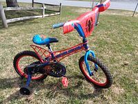 Side view of the child's Spiderman bicycle with training wheels on grassy surface showing red and blue color with Spiderman graphics.