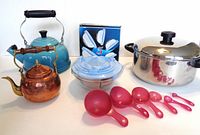 Full view showing copper kettle, blue Le Creuset kettle, unopened nested glass bowl set, stainless steel pot with lid, and nested pink measuring spoons and cups.