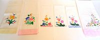 Photo showing six linen hand towels laid flat, each with a floral design embroidered or painted and some with colored borders in pastel shades of pink and peach.