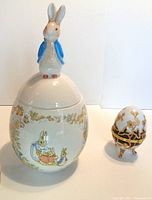 Photo showing full view of the ceramic Peter Rabbit cookie jar with attached 3D Peter Rabbit figure on lid, floral decoration on jar body, and the small decorative ceramic egg beside it.