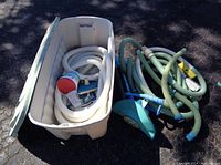 Wide view of the large beige plastic storage bin filled with coiled hoses and pool cleaning parts, next to more hoses and a blue pool cleaner attachment on the ground.
