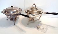 Two silverplated chafing dishes side by side on a white background. One lid is domed with black knob and is not original to its set; the other lid matches dish design with silver handle. Both dishes have ornate legs and fuel holders underneath.