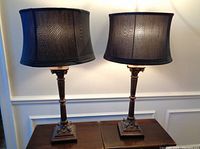 Pair of tall wooden table lamps with square bases, carved shafts, and black drum shades, placed on dark wood surface against white wall.